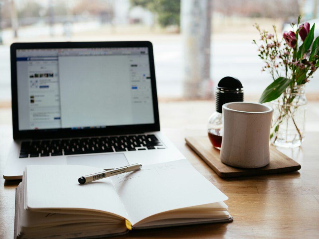 An open laptop on a table by a window with an open notebook and a cup of coffee.